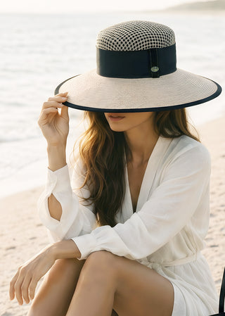 Woman wearing a handcrafted wide-brim sun hat with black band detail on a beach at sunset.