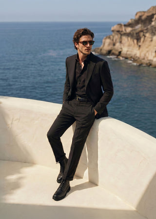 Man wearing black leather lace-up boots styled with black tailoring in a coastal setting.