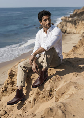 Man wearing brown leather Chelsea boots styled with relaxed tailoring on a coastal landscape.